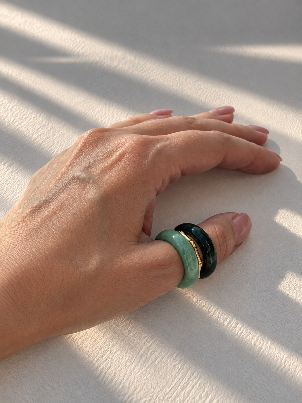 A close-up of a hand resting on a textured white surface under warm, dappled sunlight, wearing two stacked rings: one light green and one dark green with a thin gold accent.