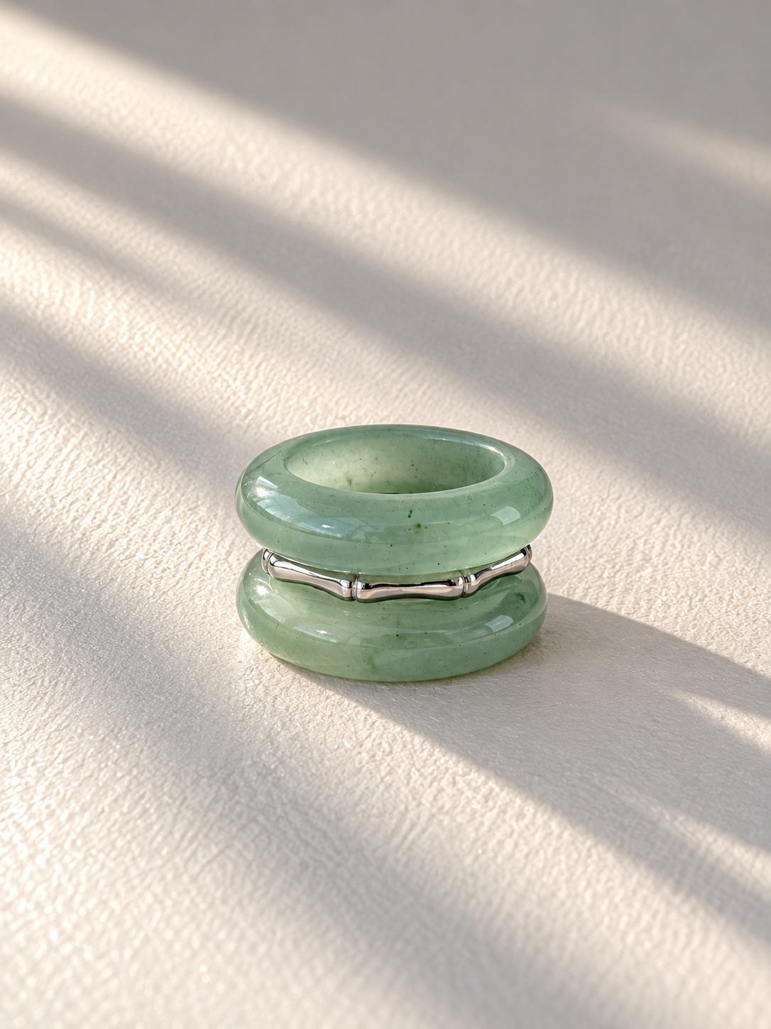 A double-band ring featuring light green jade-like stone discs stacked with a thin silver bamboo-inspired band in between, resting on a textured cream surface with soft diagonal shadows.