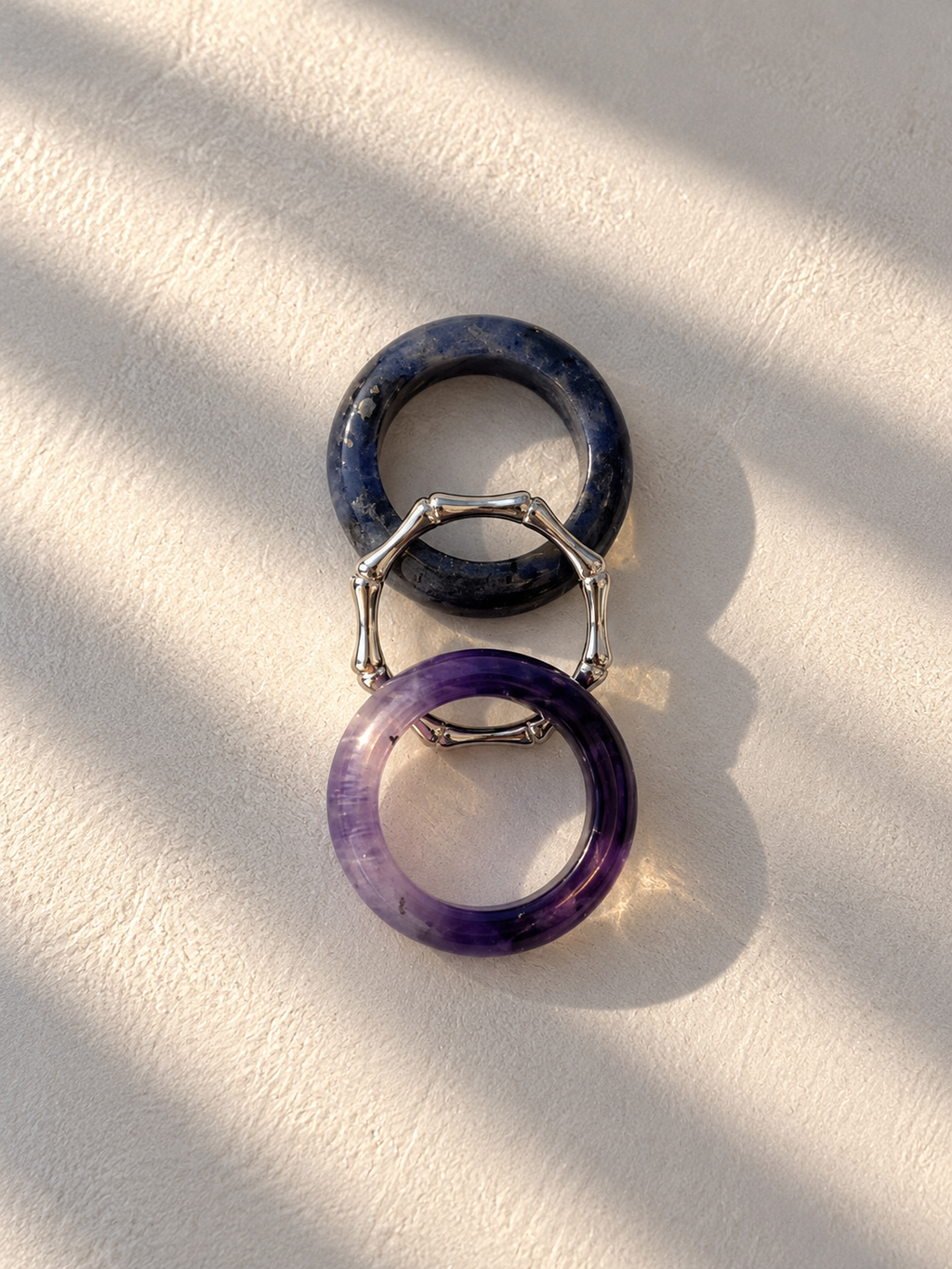 Two circular ring-shaped earrings or charms, one dark navy blue and one purple, connected by a silver-toned geometric bridge, displayed on a textured off-white surface with diagonal shadows.