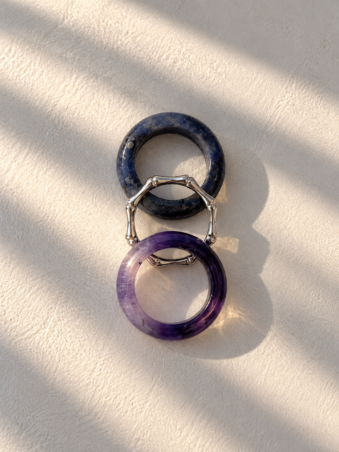 Two circular ring-shaped earrings or charms, one dark navy blue and one purple, connected by a silver-toned geometric bridge, displayed on a textured off-white surface with diagonal shadows.