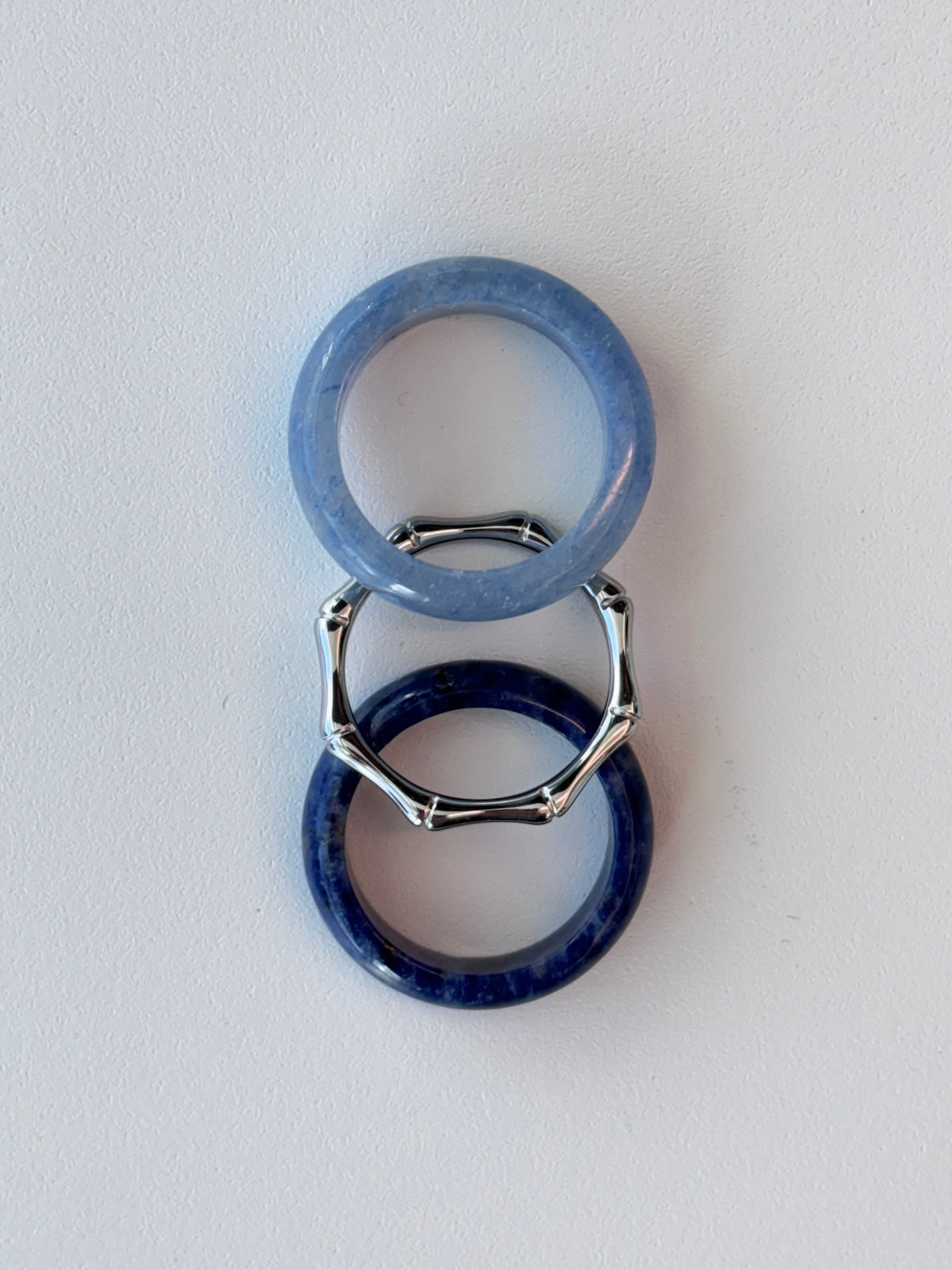 Three rings stacked vertically on a white background: a light blue stone ring at the top, a silver-toned geometric ring in the middle, and a dark navy blue stone ring at the bottom.