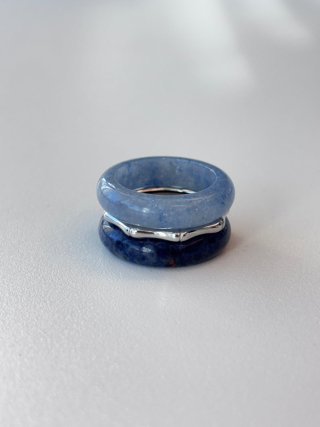 A stack of three rings on a light gray surface, featuring a light blue stone band on top, a dark blue stone band on the bottom, and a silver-toned metal band in the middle.