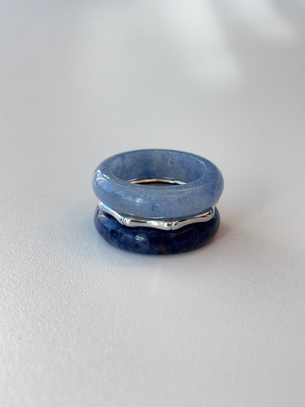 A stack of three rings on a light gray surface, featuring a light blue stone band on top, a dark blue stone band on the bottom, and a silver-toned metal band in the middle.