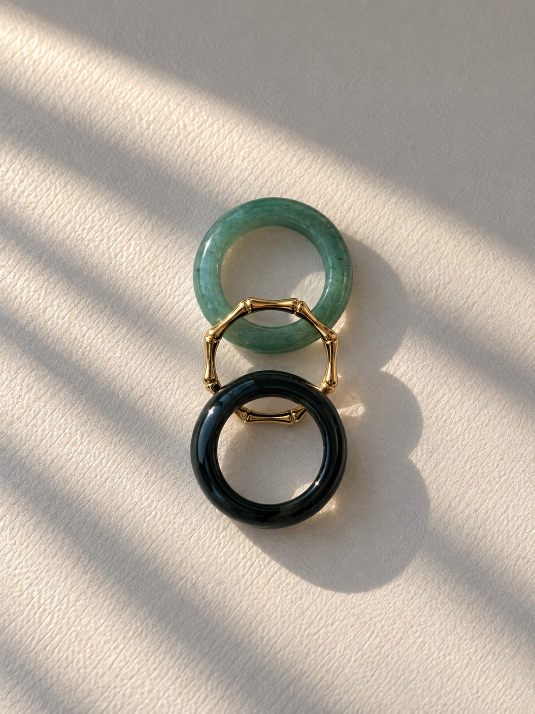 Two geometric ring earrings with gold-toned metal and a green translucent top ring and a black opaque bottom ring, displayed on a textured off-white surface with diagonal shadows.