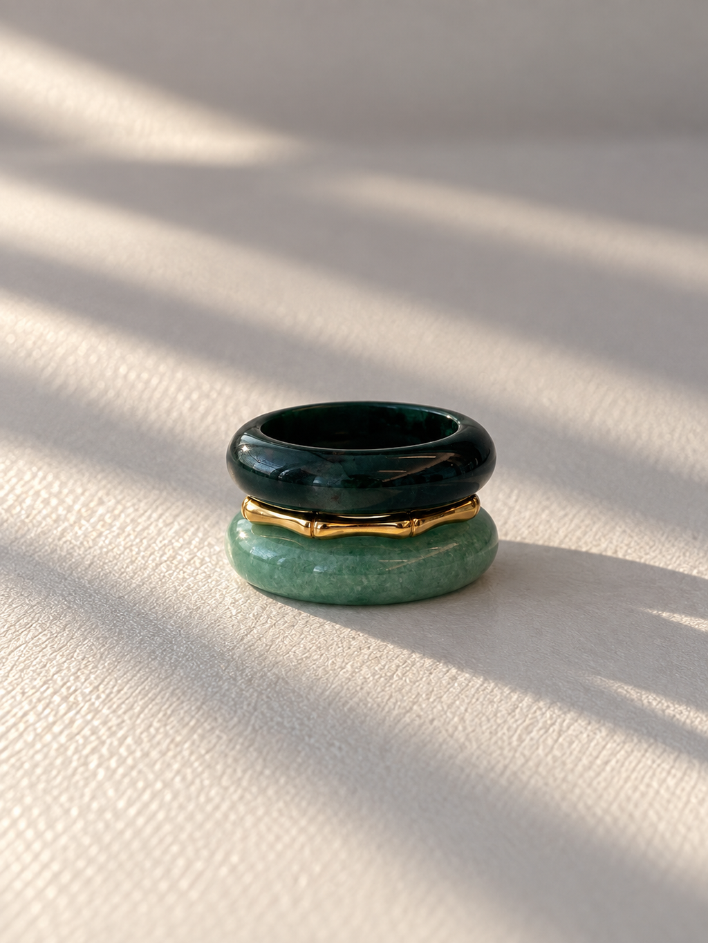 A stack of three rings on a textured beige surface with soft diagonal shadows. The top ring is dark green polished stone, the middle ring is a thin gold metal band, and the bottom ring is a chunky light green stone.