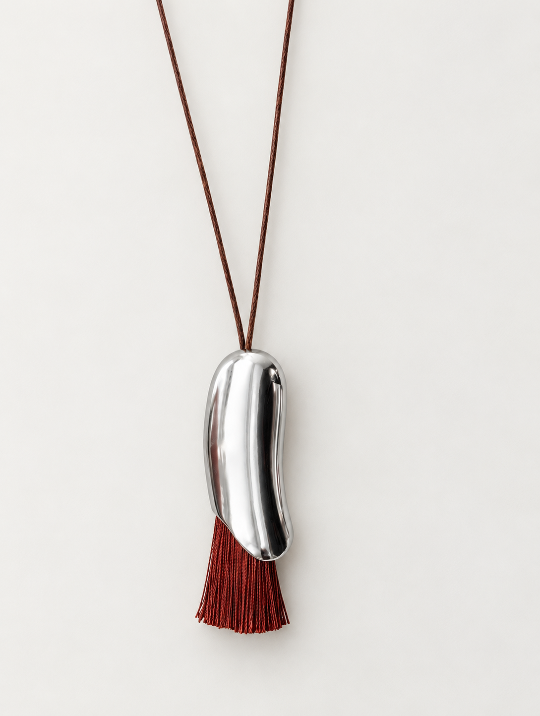 A minimalist shiny pendant necklace featuring a polished silver bean-shaped charm suspended on a dark brown cord, with a matching dark red brown tassel hanging below.