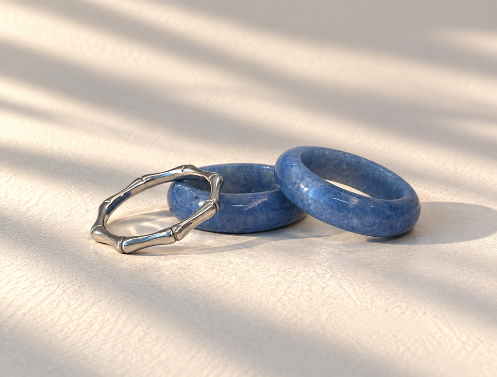 Two rings on a textured cream surface with soft leaf shadows: one is a polished silver-toned ring with a segmented bamboo-like design, and the other is a thick blue stone ring. No visible brand markings.