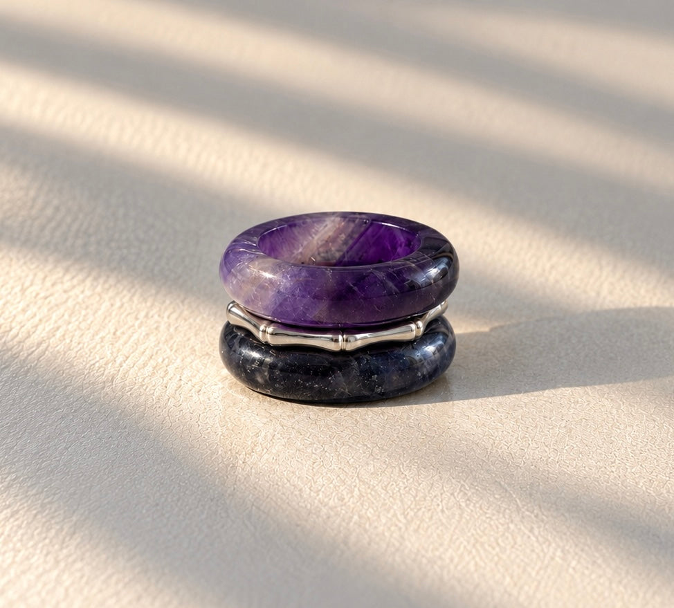 A two-stone ring featuring a polished purple amethyst top stone and a dark navy blue lapis lazuli bottom stone, separated by a silver-toned bamboo band, resting on a textured cream surface with soft diagonal shadows.
