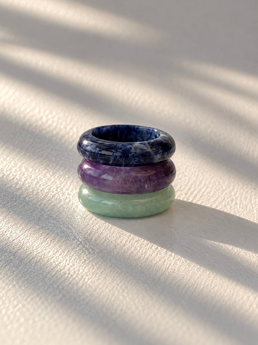 Three stacked bangle rings made of polished stone, featuring a dark blue top ring, a purple middle ring, and a light green bottom ring, displayed on a textured cream surface with soft diagonal shadows.