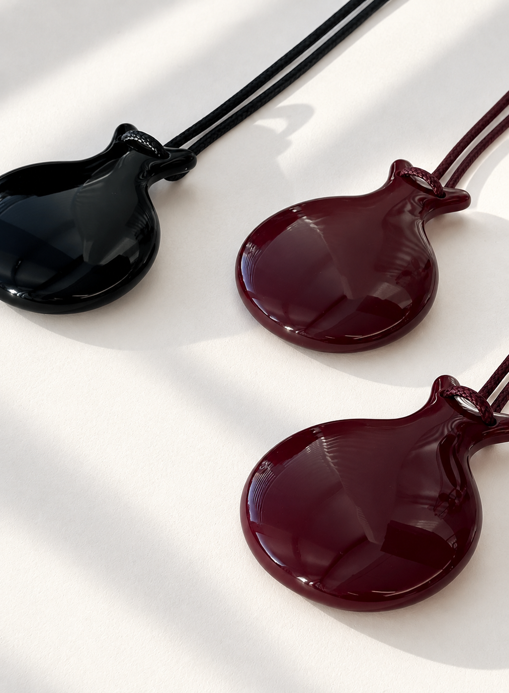Three glossy round castanet-shaped pendant necklaces on cords, shown on a white surface with soft shadows. pendants are dark burgundy, brown and black.