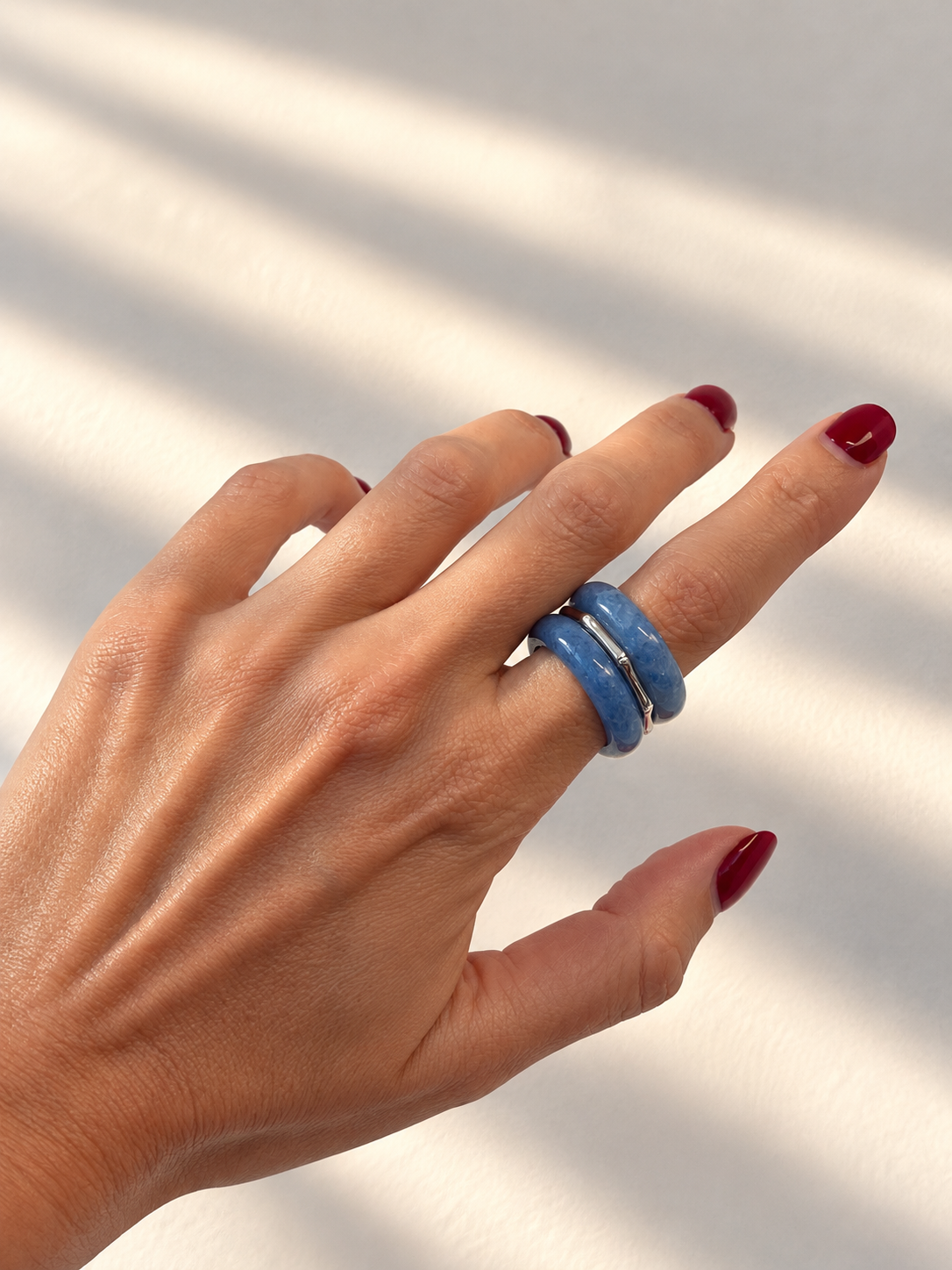 A close-up of a hand with dark red nail polish wearing two stacked blue rings against a white background with soft diagonal shadows.