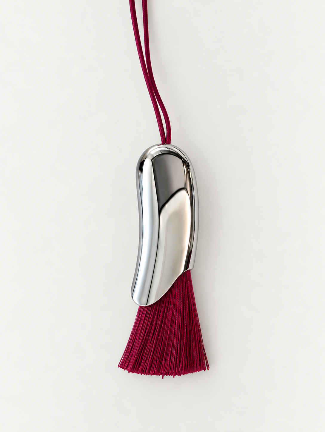 A polished silver-toned metal pendant shaped like a bean or abstract organic form, hanging from a deep magenta cord with a matching tassel at the bottom, set against a clean off-white background.