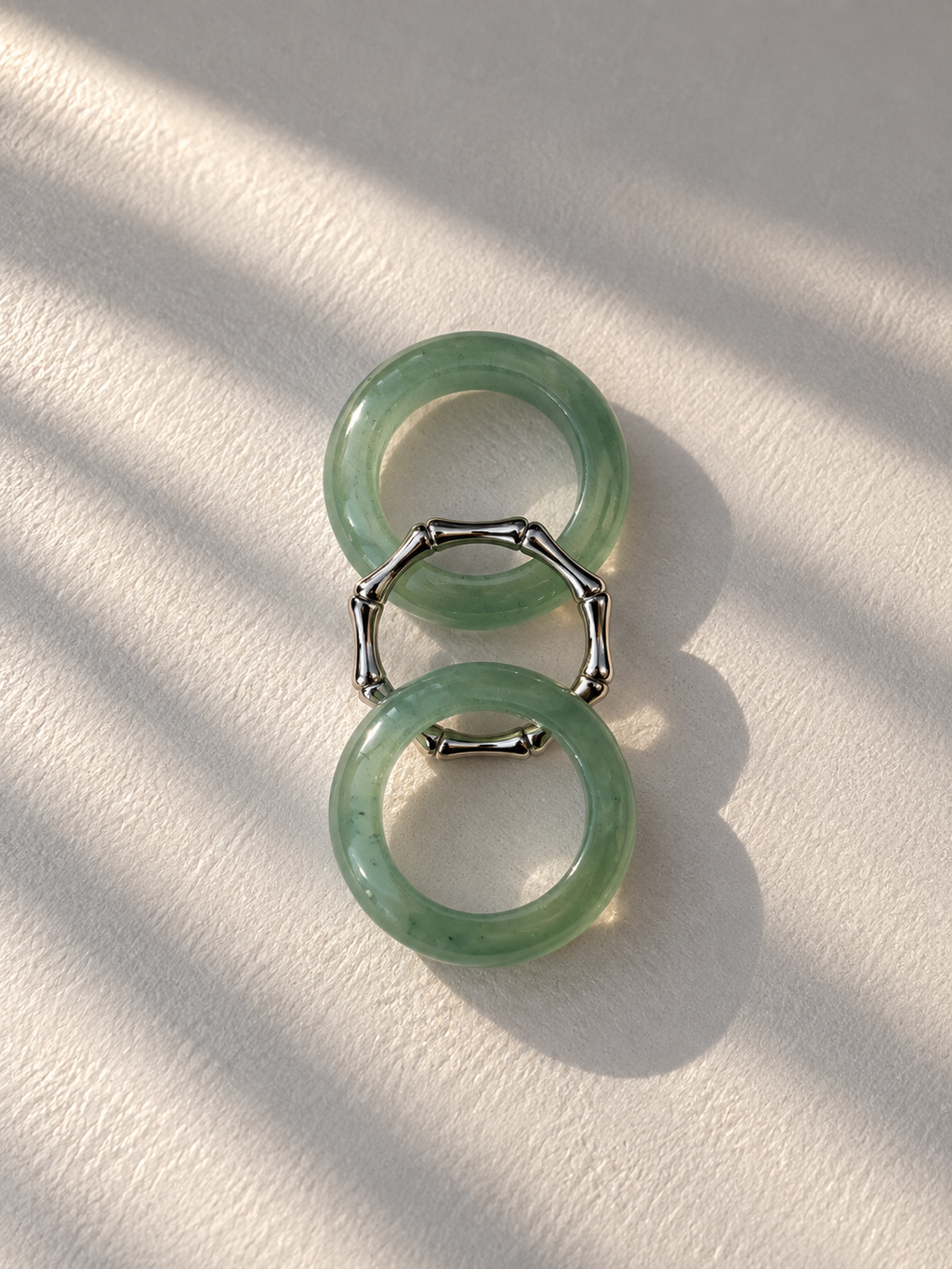 A pair of green jadeite bangle rings connected by a silver-toned bamboo-style ring, displayed on a textured off-white surface with soft diagonal shadows.