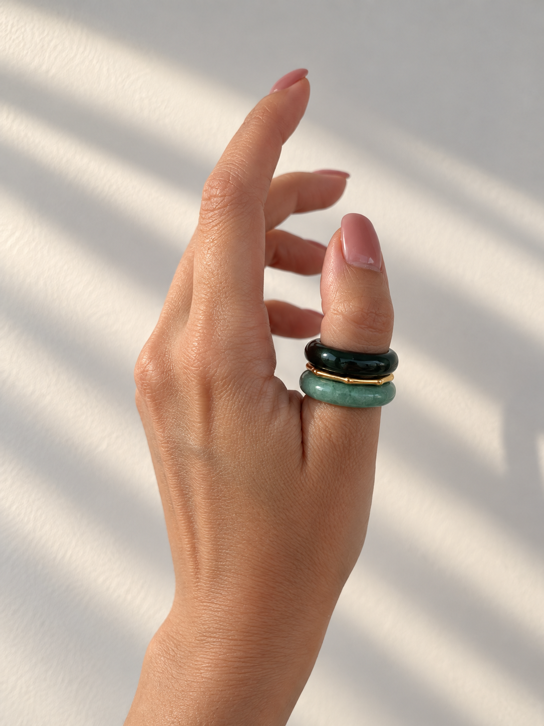 A close-up of a hand wearing three stacked ring bracelets, featuring a dark green stone, a black stone with a gold band, and a light green stone, set against a warm off-white background with soft leaf shadows.