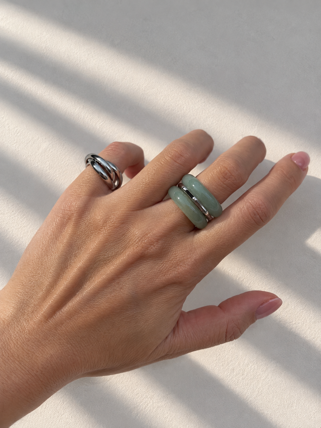 A close-up of a hand wearing two rings: a silver wrap-style band on the ring finger and a light green jade-like band on the middle finger, set against a sunlit off-white background with soft shadows.