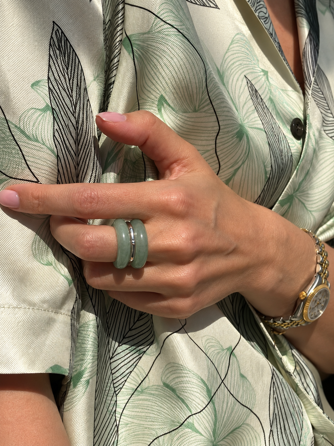 A close-up of a person's hand wearing two green jadeite rings, one larger and one smaller, with a gold and silver watch on the wrist, wearing a green and beige patterned shirt.