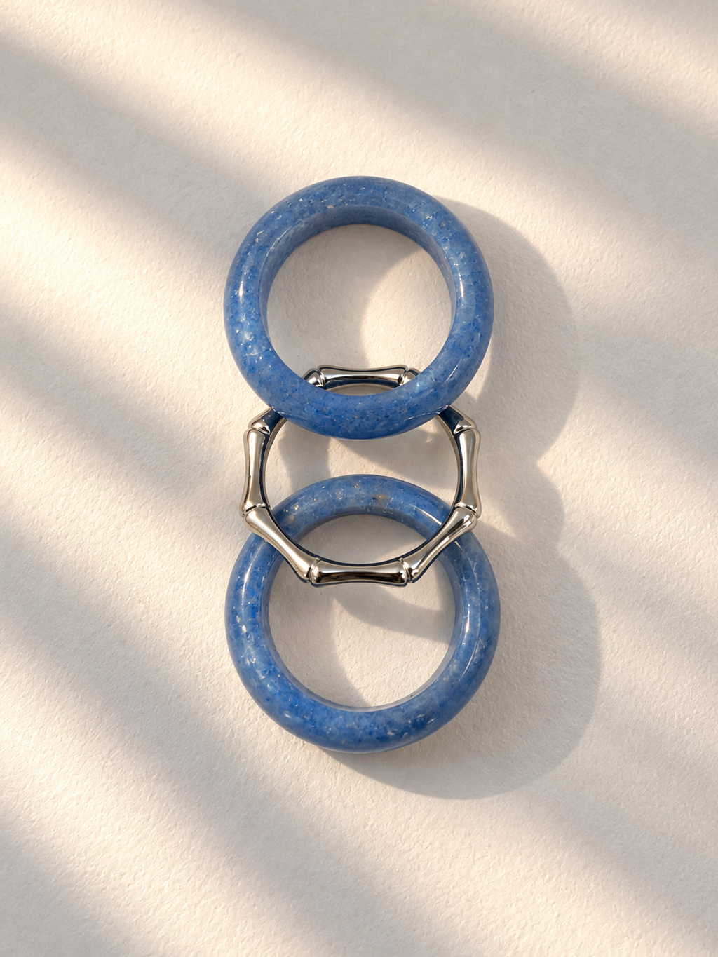 Two blue stone rings connected by silver-toned bamboo-style links, photographed on a warm off-white surface with soft diagonal shadows.