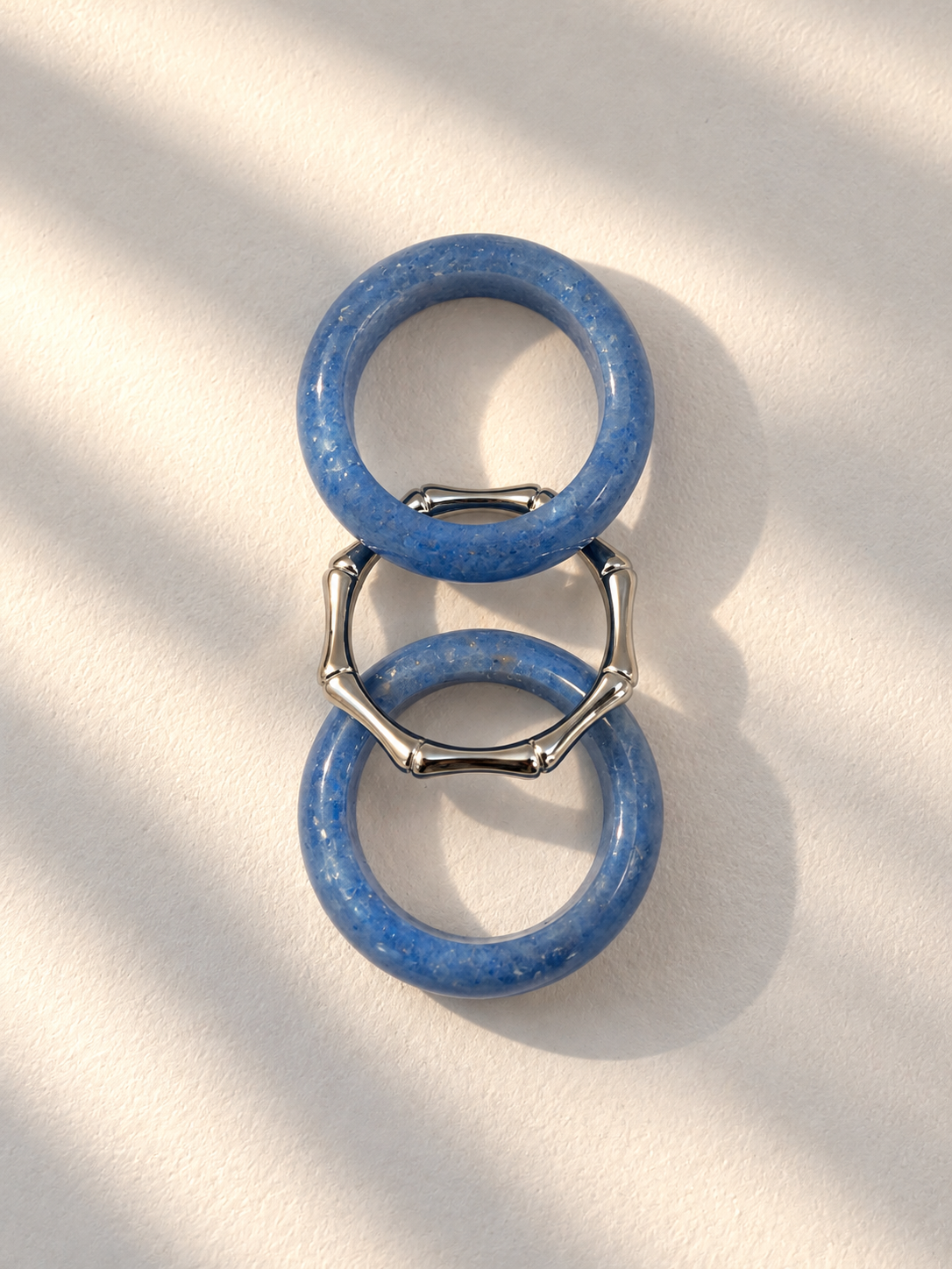 Two blue stone rings connected by silver-toned bamboo-style links, photographed on a warm off-white surface with soft diagonal shadows.