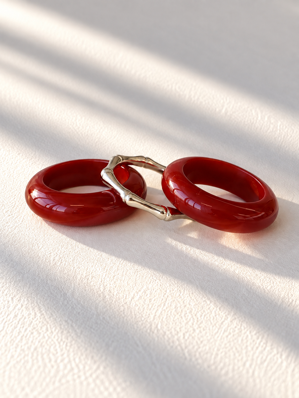 Two glossy red circular earrings connected by a silver-toned bamboo-style bridge, photographed on a white textured surface with soft diagonal shadows.