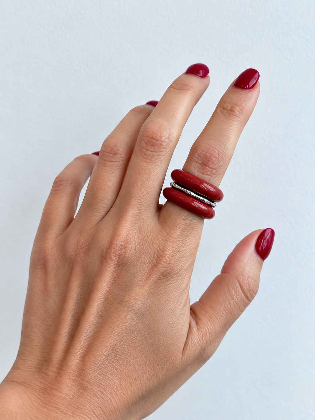 A close-up of a hand with dark red manicured nails wearing a chunky, multi-band ring made of glossy dark red cylindrical elements.
