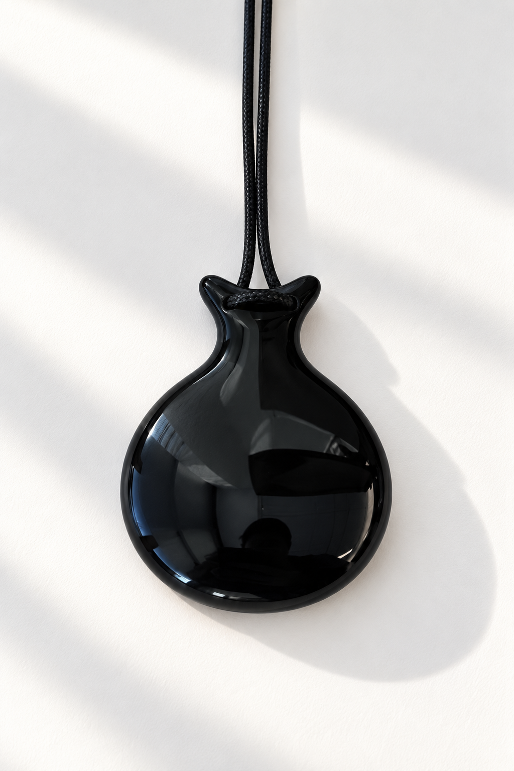 A glossy black pendant shaped like a small teardrop or money bag, suspended from a black cord against a bright white background with soft diagonal shadows.