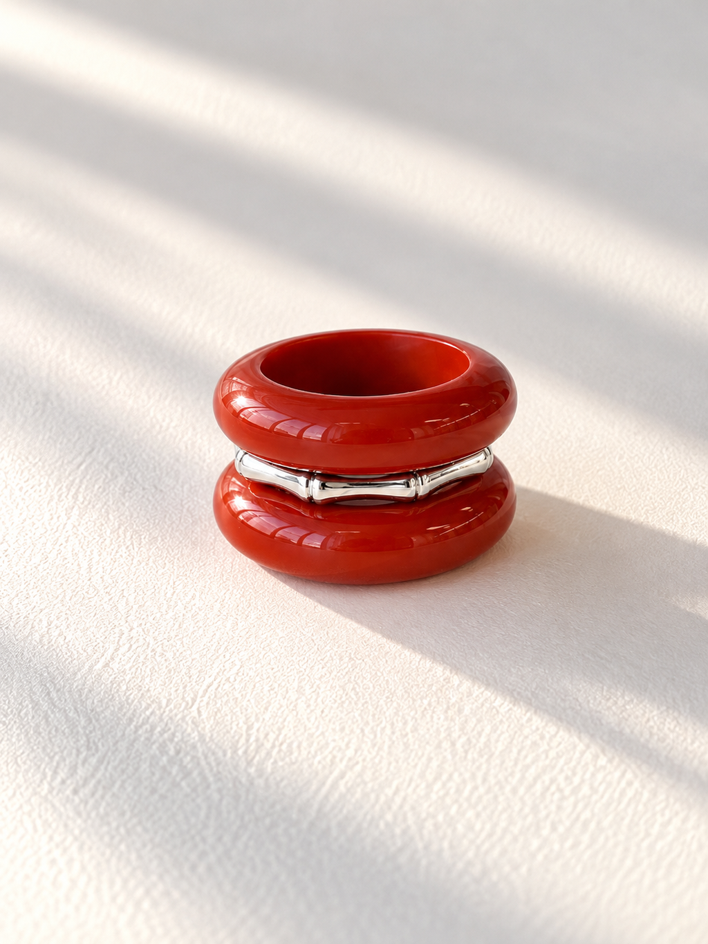 A glossy red double-ring ring with a silver bamboo-style band in the middle, photographed on a textured white surface with soft diagonal shadows.
