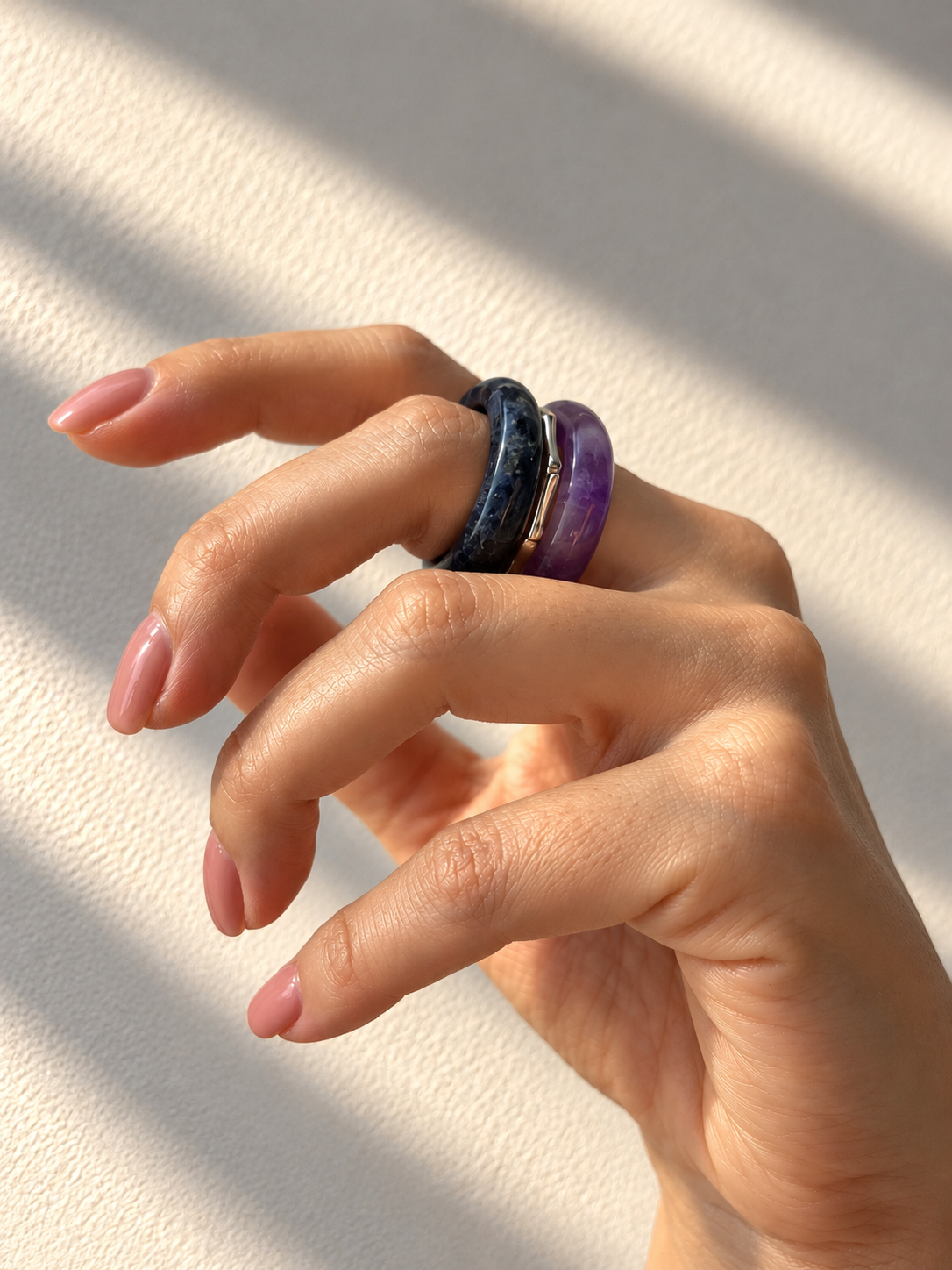 A close-up of a hand wearing two stacked rings, one dark blue with white speckles and one purple, set against a textured off-white background with soft diagonal shadows.