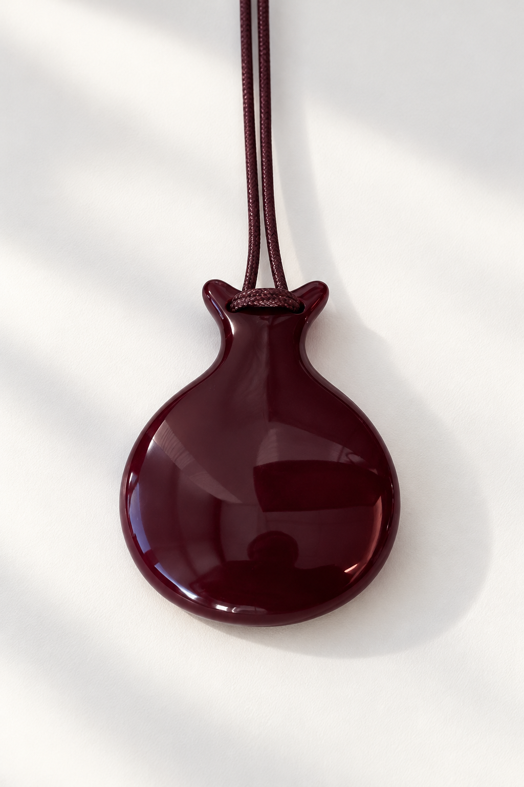 A glossy dark burgundy pendant shaped like a castanet, suspended from a matching dark cord against a white background with soft diagonal shadows.
