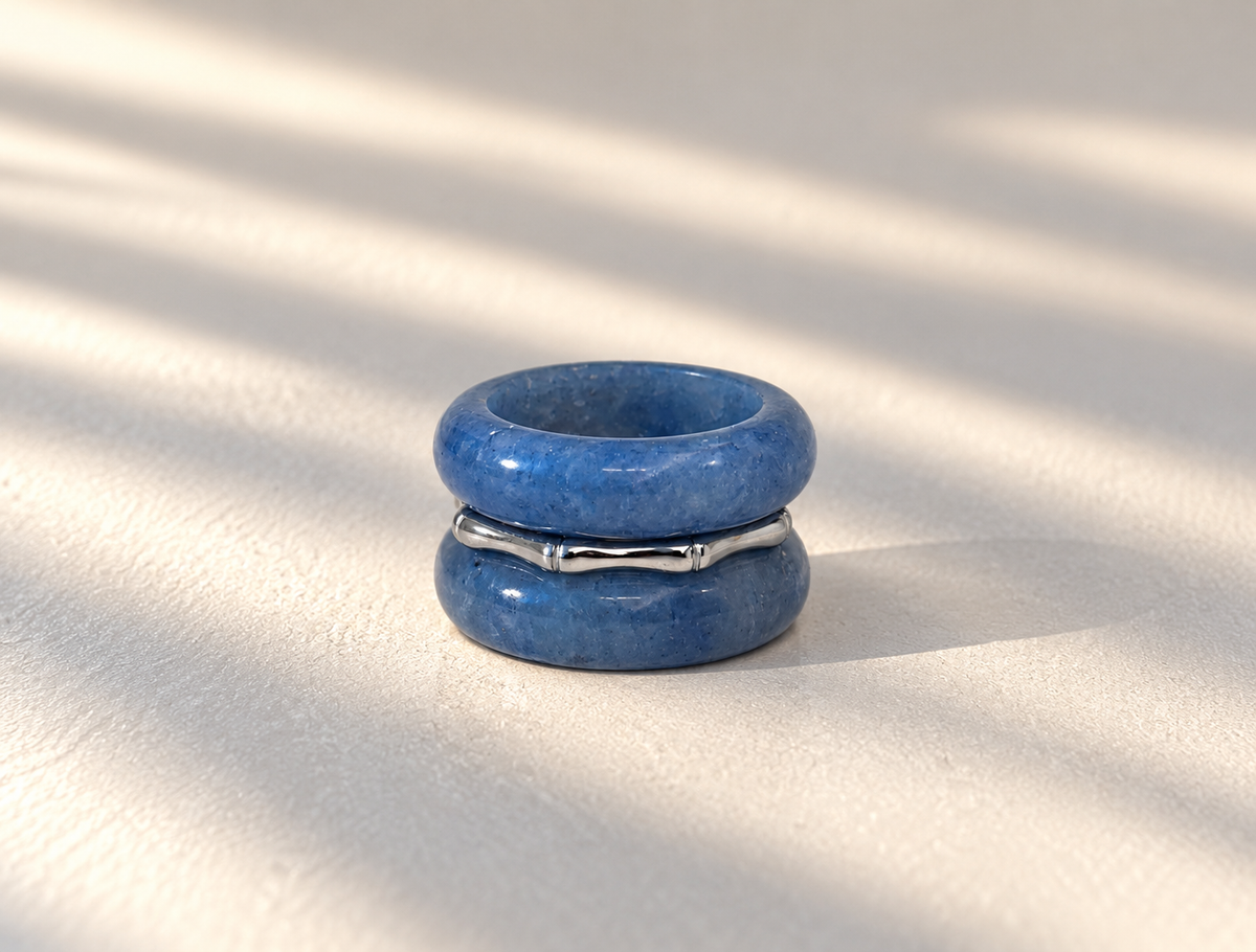A pair of stacked blue stone rings with a polished silver band in between, photographed on a light textured surface with soft diagonal shadows.