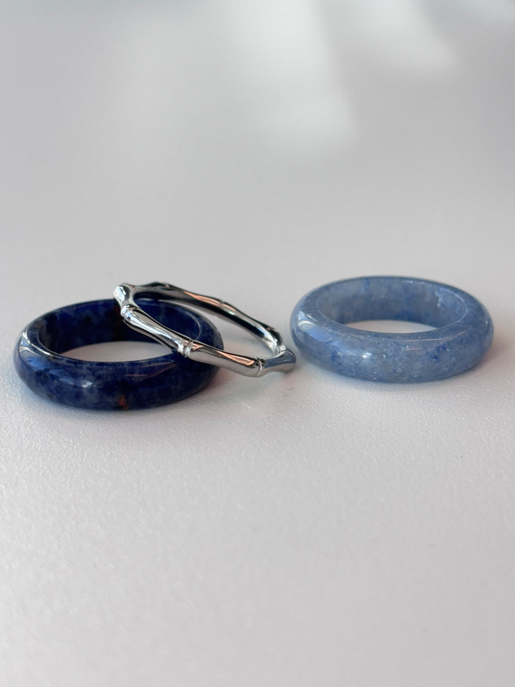 Three rings displayed on a light gray surface: a dark blue stone ring, a lighter blue stone ring, and a silver-toned twisted ring resting on top of the dark blue ring.