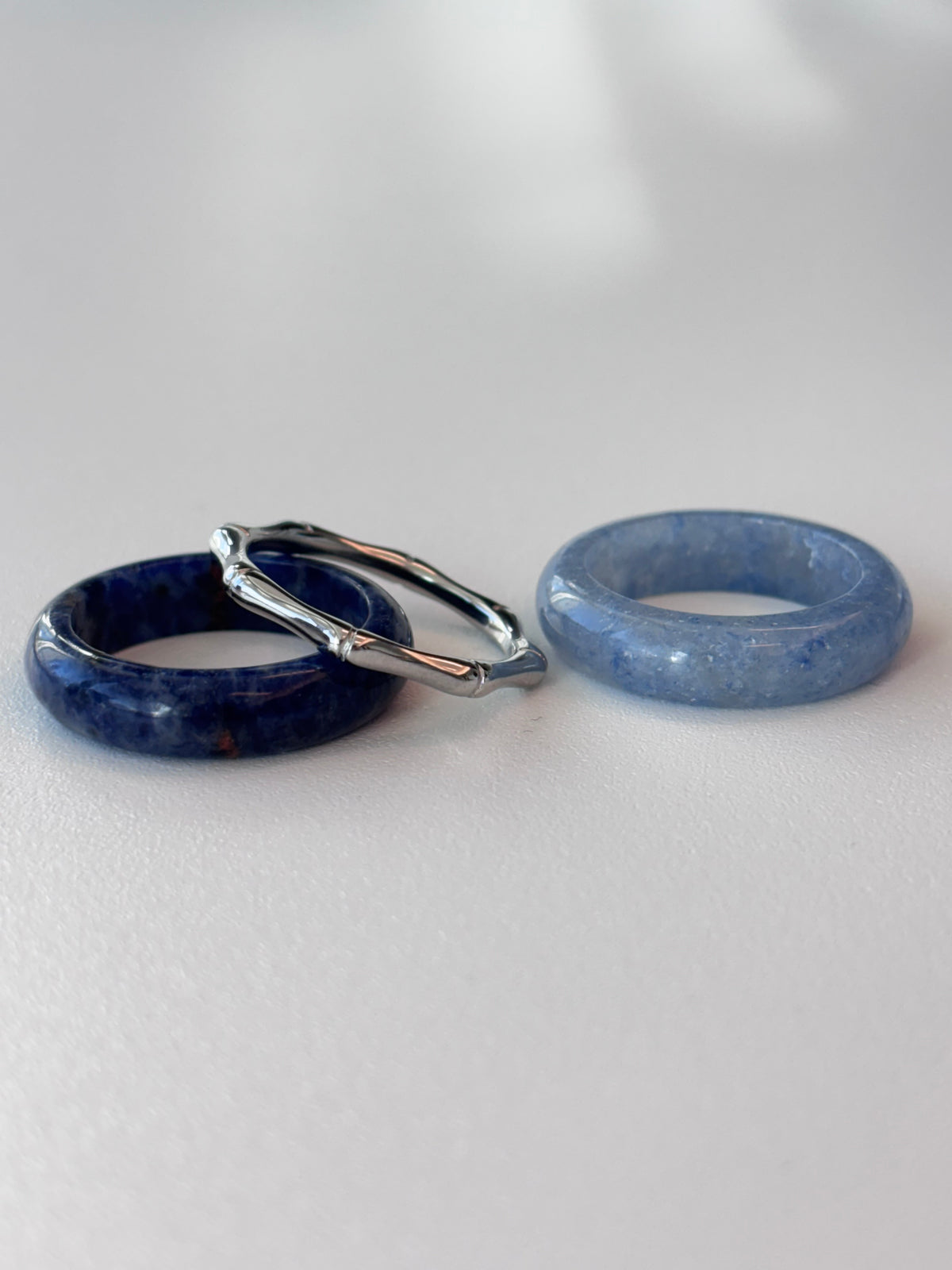 Three rings displayed on a light gray surface: a dark blue stone ring, a lighter blue stone ring, and a silver-toned twisted ring resting on top of the dark blue ring.