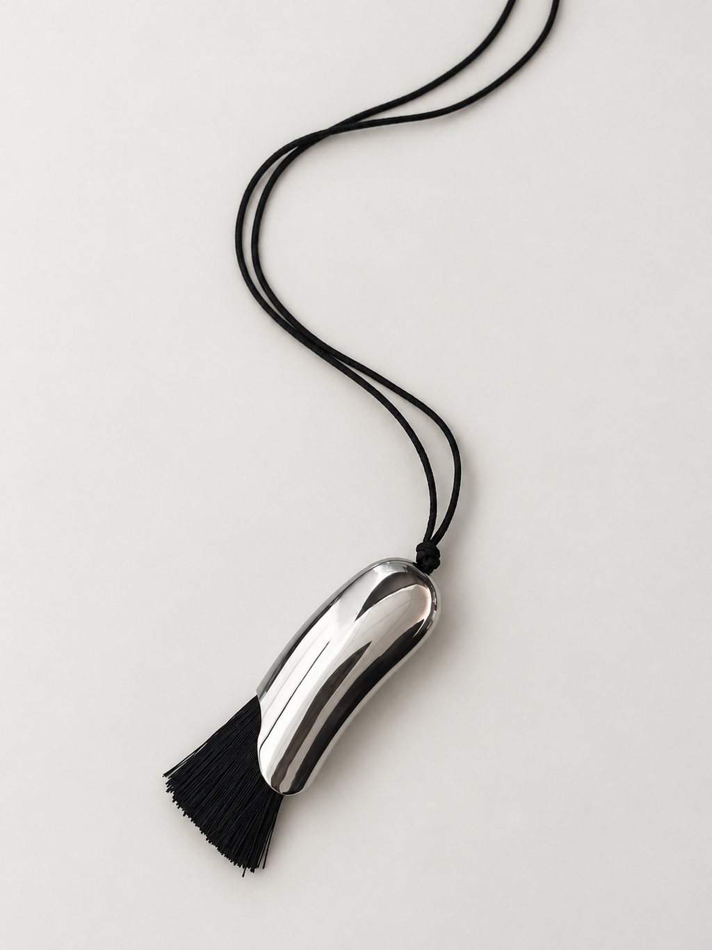 A modern silver metallic pendant necklace with a black tassel and black cord, photographed on a clean, light gray background.