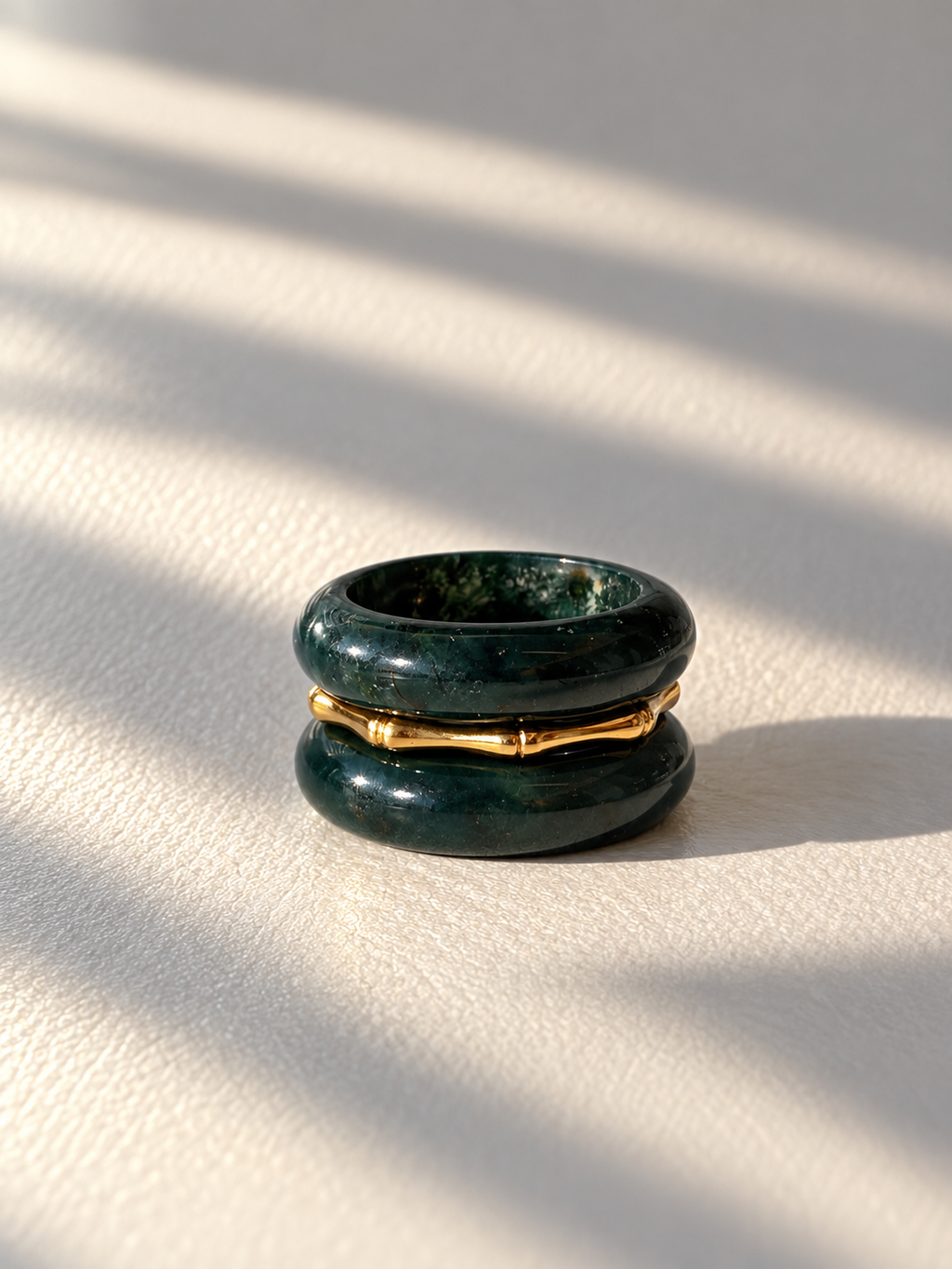 A double-band ring featuring dark green stone inlays and a polished gold metallic center band, resting on a textured cream surface with soft diagonal shadows.
