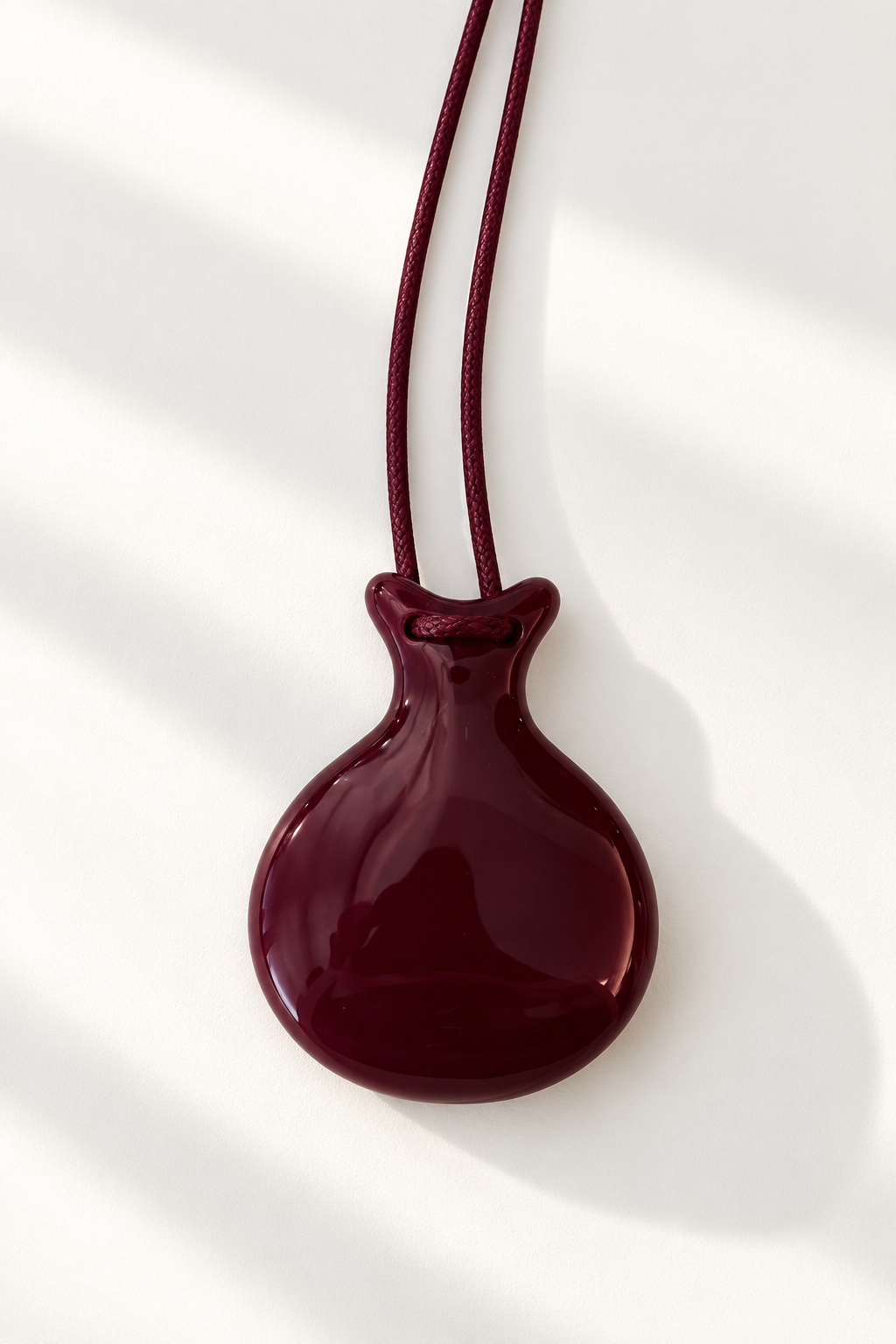 A glossy dark red pendant shaped like a small bag or teardrop, suspended from a matching dark red cord against a white background with soft diagonal shadows.