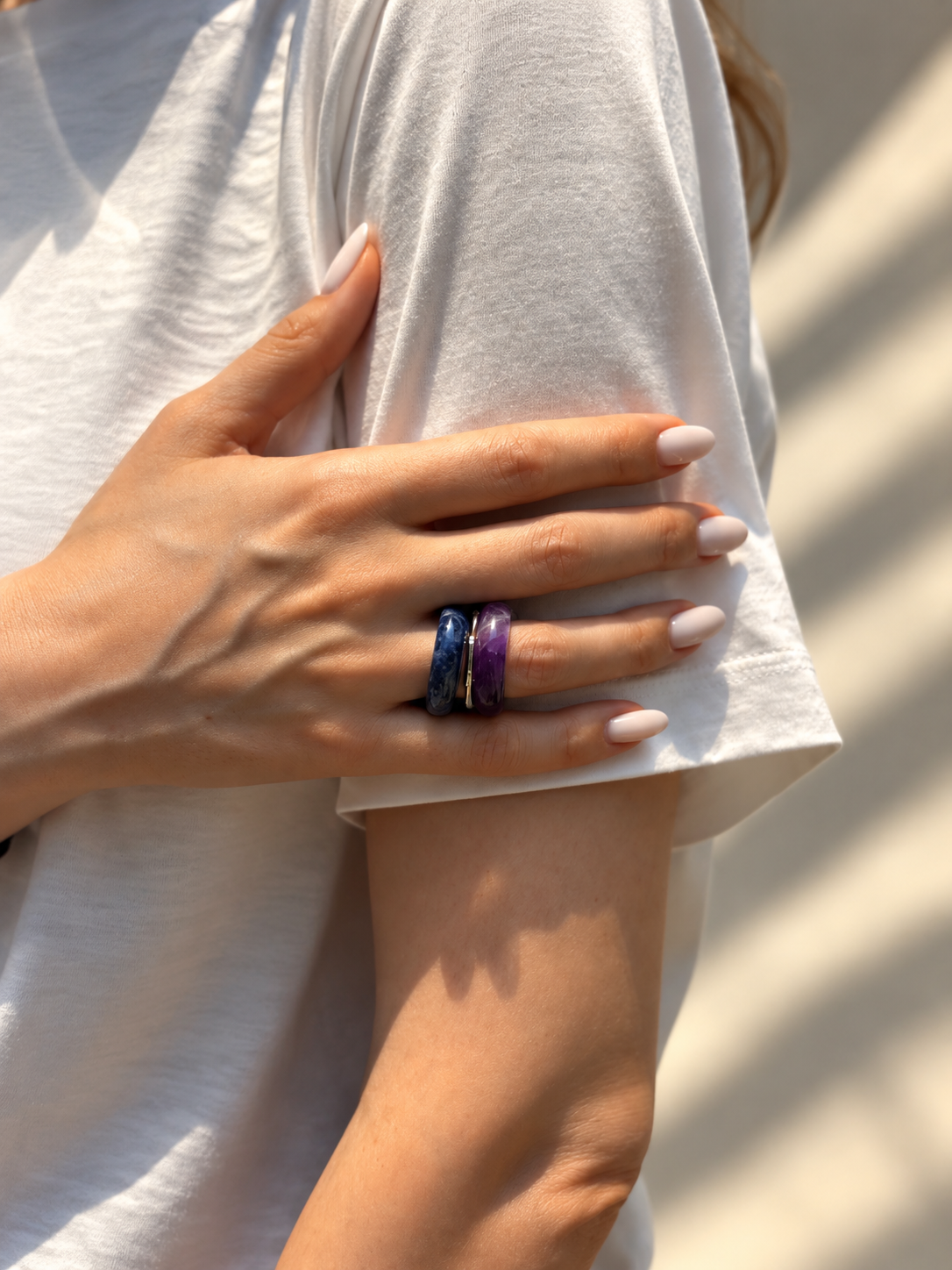 A close-up of a person's hand resting on their arm, wearing two stacked rings: one dark blue with a starry pattern and one deep purple, both with thin gold edges. The person is wearing a white t-shirt in bright natural light.