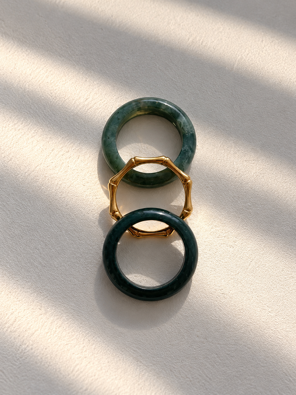 Three rings arranged vertically on a textured off-white surface with soft diagonal shadows. The top and bottom rings are dark green stone with gold-toned metal accents, and the center ring is a hexagonal gold-toned metal ring.