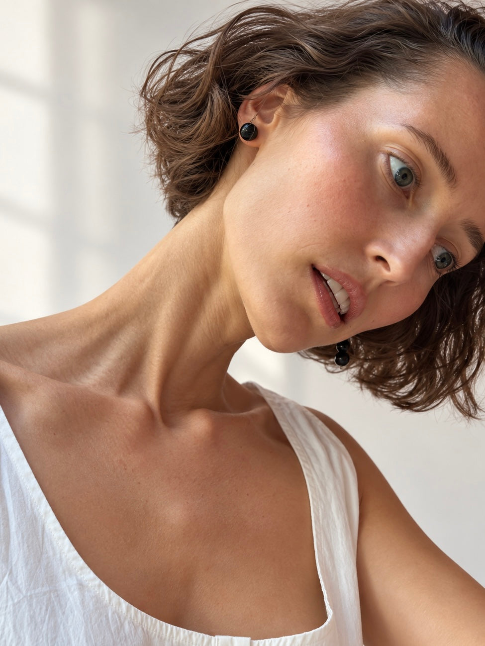 A close-up lifestyle shot of a woman with short brown hair wearing small black round stud earrings and a white top, looking down and to the side in a brightly lit indoor setting.