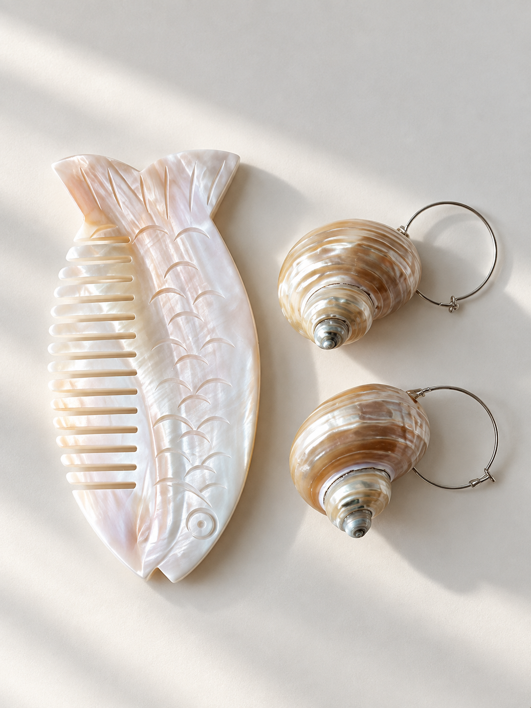 A set of three shell-inspired jewelry accessories arranged on a light beige surface with soft diagonal shadows: a large fish-shaped comb on the left, and two spiral shell-shaped hoop earrings on the right.