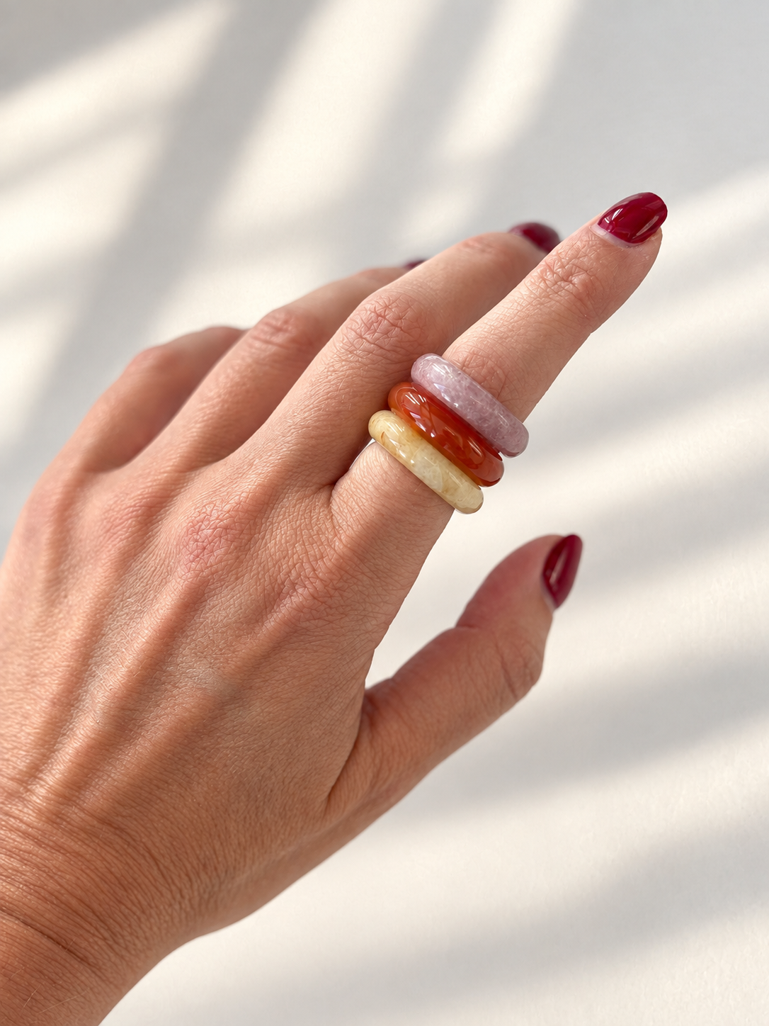 A close-up of a hand with dark red nail polish wearing three stacked, smooth stone rings in pale pink, amber orange, and cream yellow, set against a softly lit white background with natural shadow patterns.
