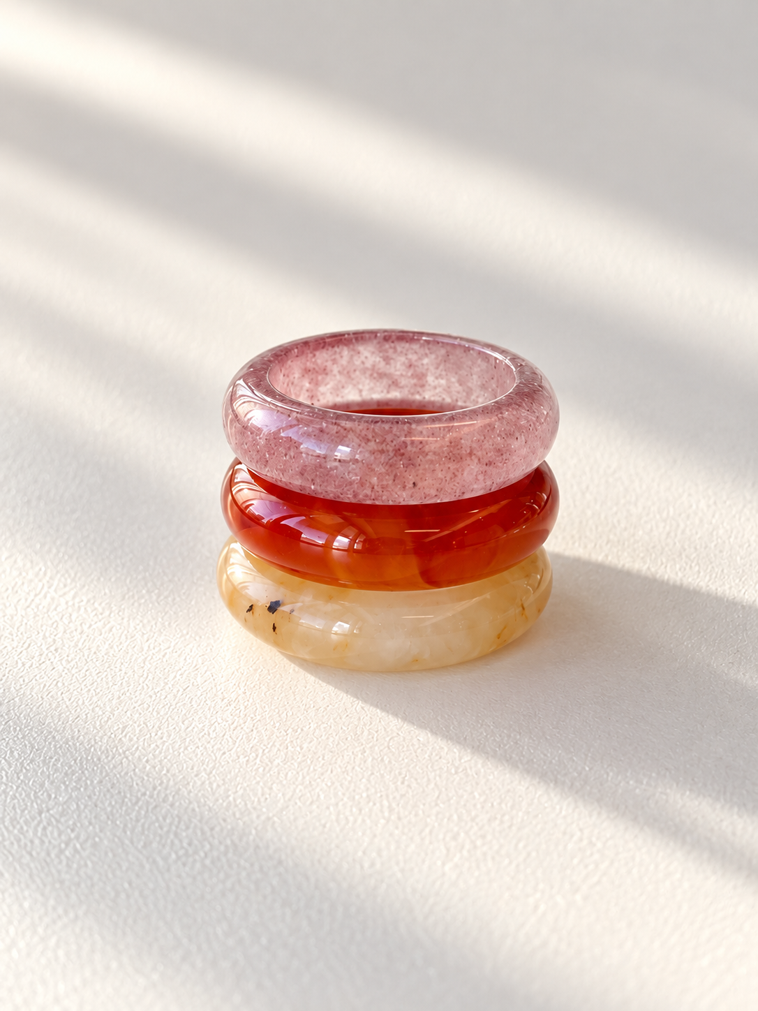 Three stacked polished stone rings in pinkish-red, orange-red, and pale yellow tones, photographed on a light textured surface with soft diagonal shadows.