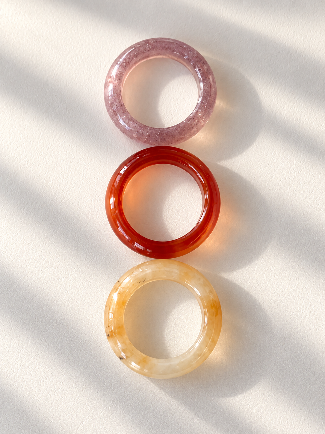 Three round translucent bangles stacked vertically on a white surface with soft diagonal shadows. The top bangle is light pink, the middle is translucent orange-red, and the bottom is translucent pale yellow with natural marbling.