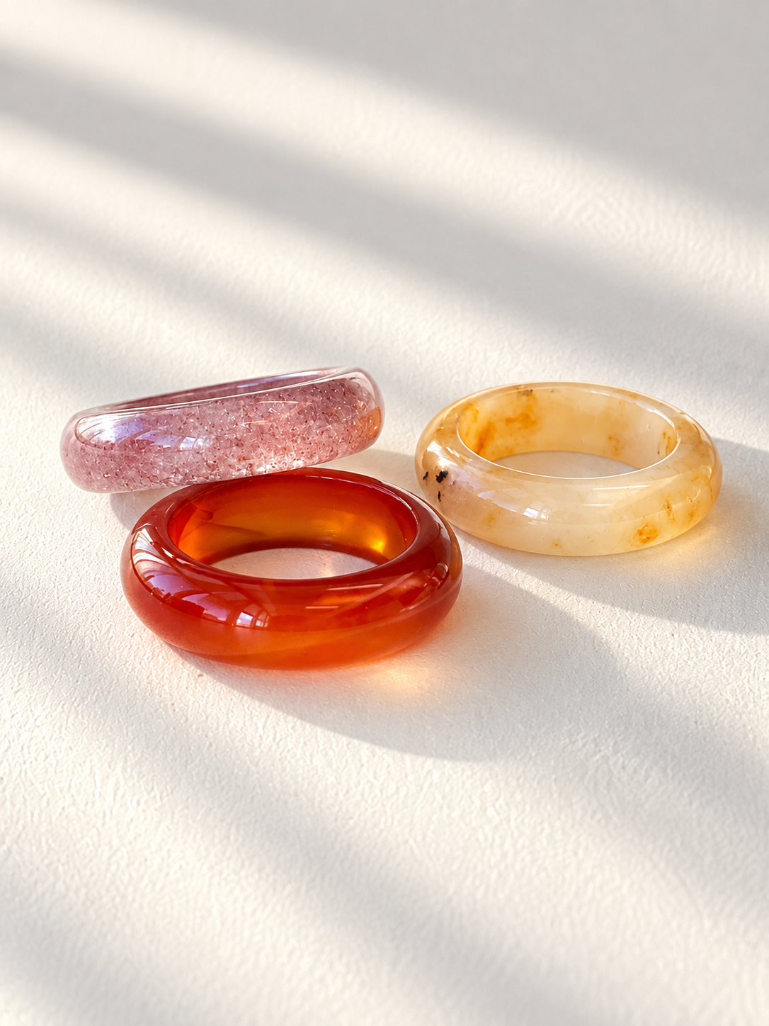 Three translucent ring-shaped bangles in pink, amber red, and cream-yellow with natural marbling, arranged on a textured white surface with soft diagonal shadows.
