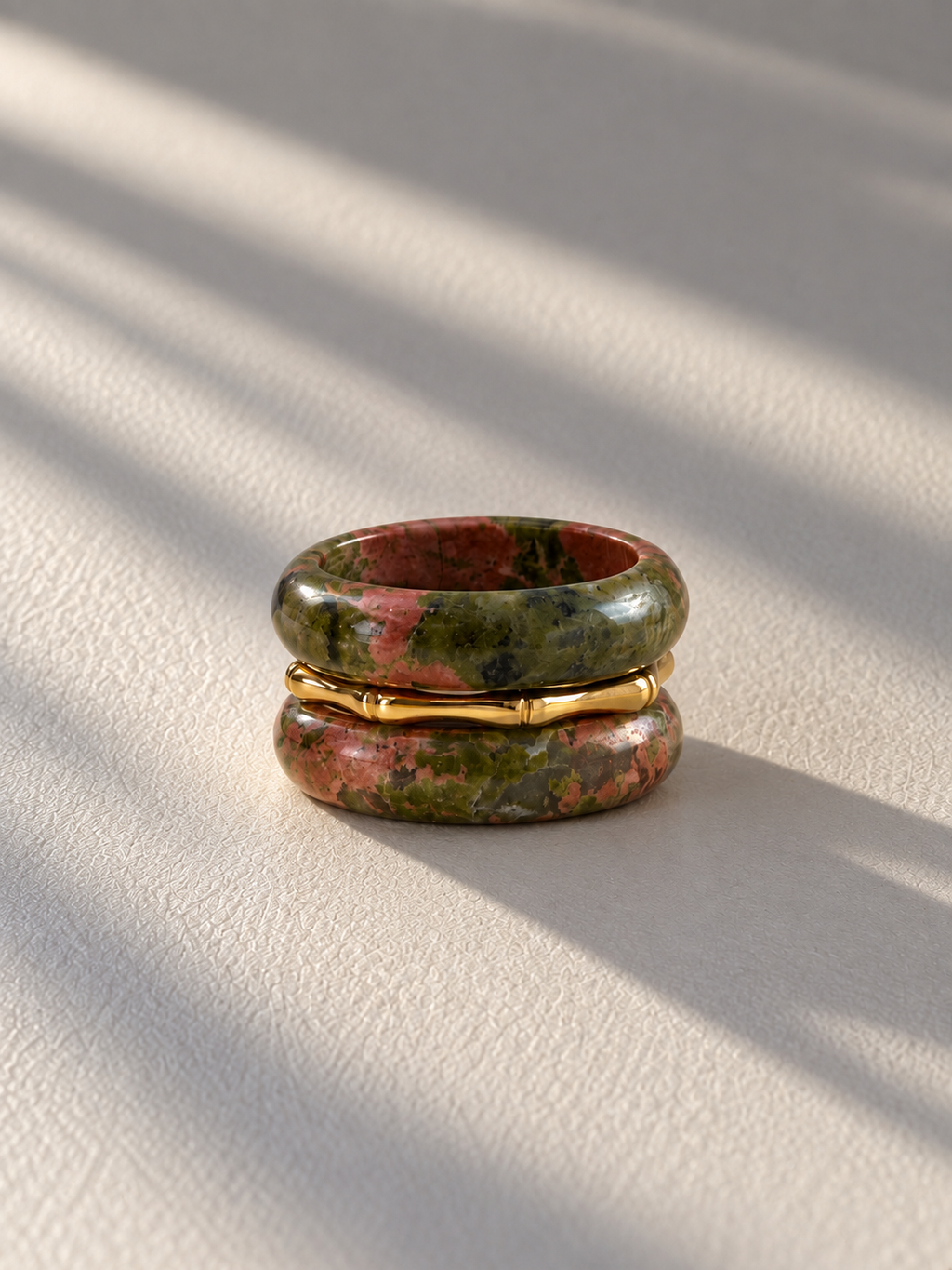 A pair of stacked rings on a textured cream surface with soft diagonal shadows. The top and bottom rings are made of a green and brown marbled stone, and the center ring is a thin gold band.