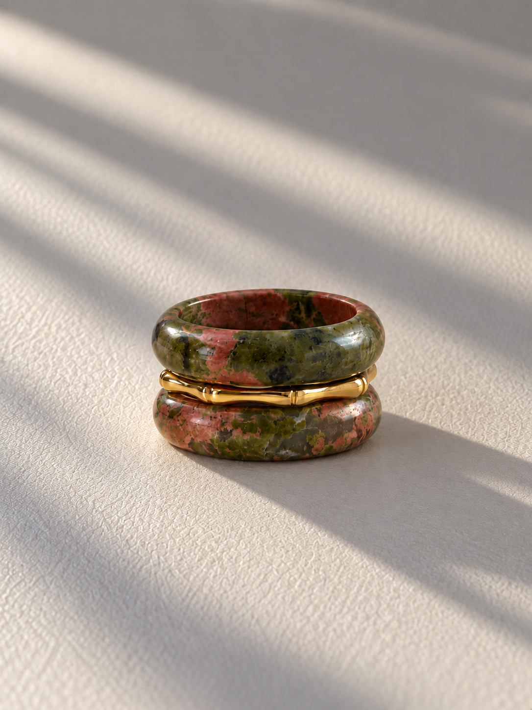 A pair of stacked rings on a textured cream surface with soft diagonal shadows. The top and bottom rings are made of a green and brown marbled stone, and the center ring is a thin gold band.