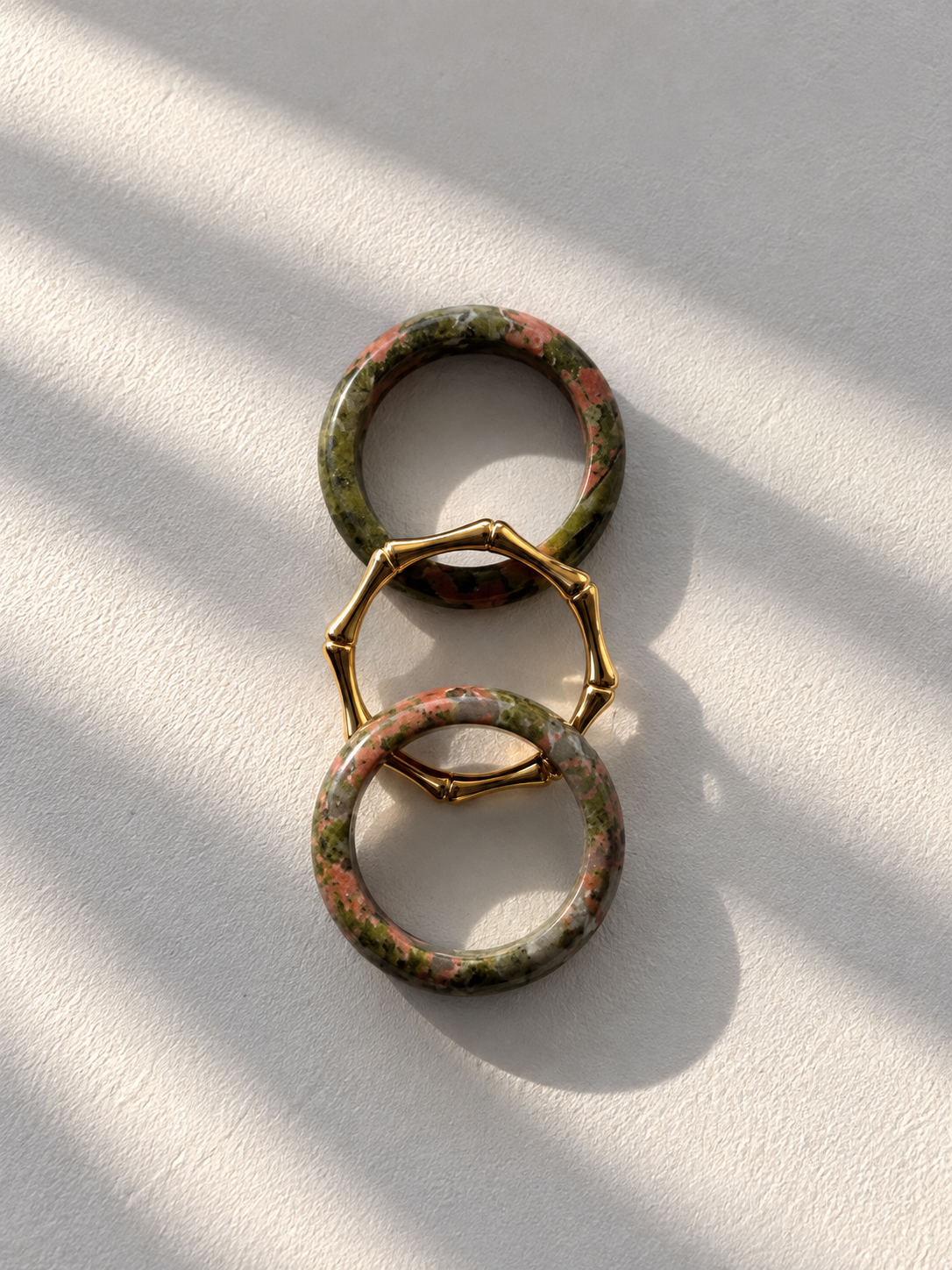 Three stacked geometric rings with a green, brown marbled pattern and gold-toned metal accents, displayed on a textured white surface with soft diagonal shadows.