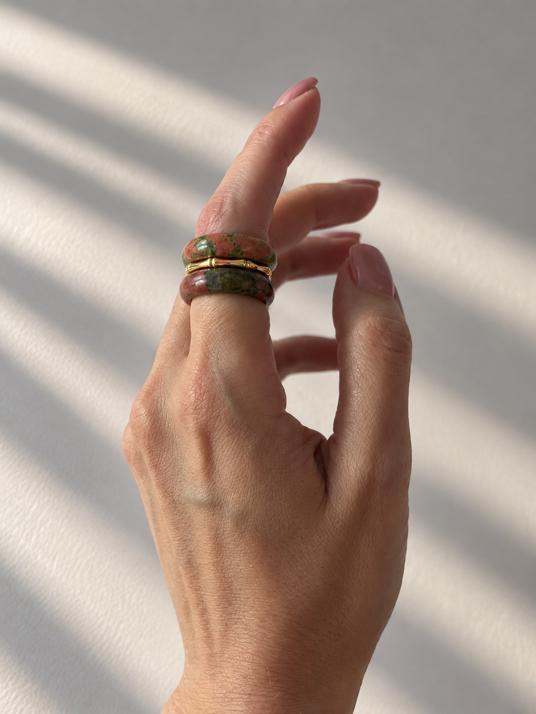 A close-up of a hand wearing three stacked rings, made of green and red marbled stone and the other a thin gold band, set against a white background with soft diagonal shadows.