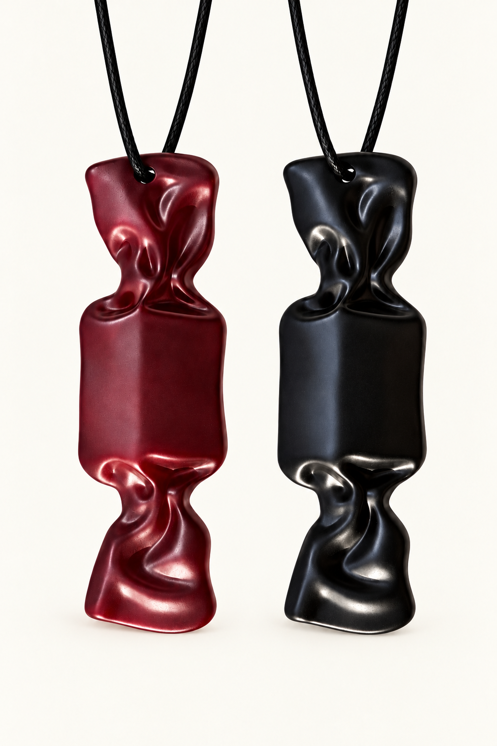 Two sculptural pendant necklaces with abstract candy, crumpled forms hanging from black cords, shown on a plain light background. One pendant is deep red and the other is black.