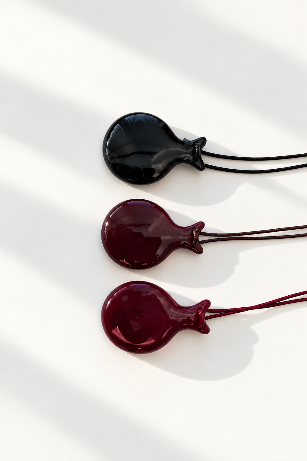 Three round castanet-shaped pendant necklaces in glossy black, deep burgundy, and brown, arranged vertically on a white background with soft diagonal shadows.
